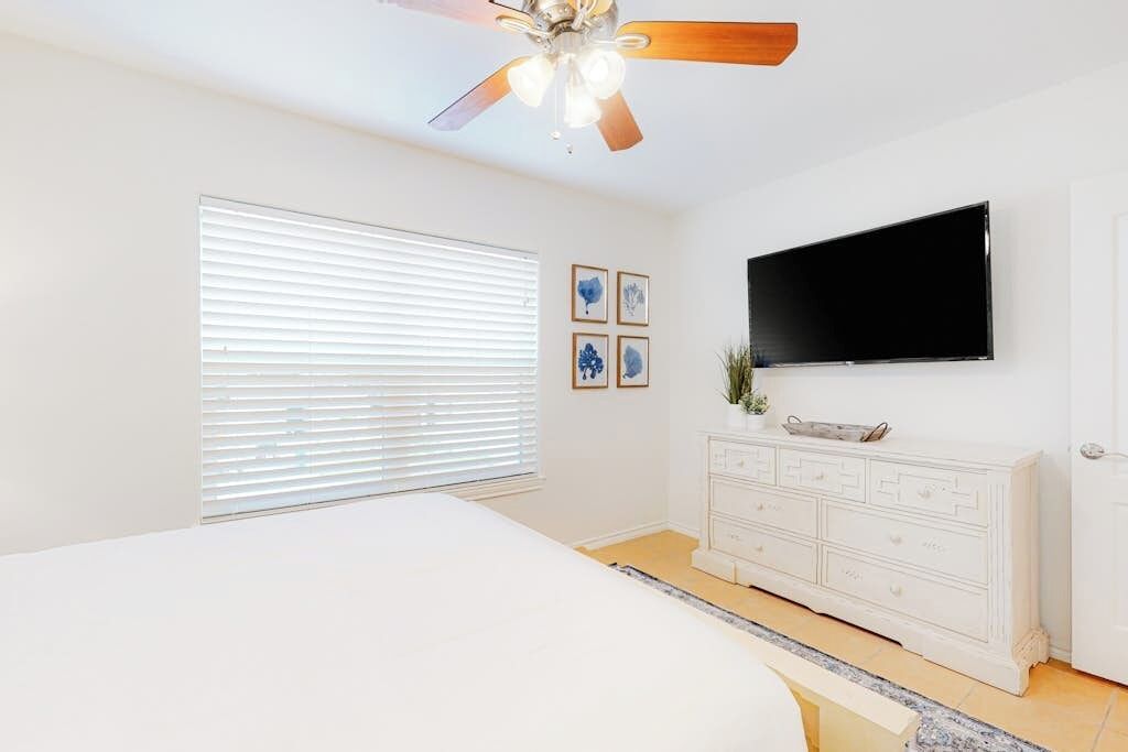 A bedroom with a bed, a ceiling fan, a TV mounted on the wall, and a white dresser.