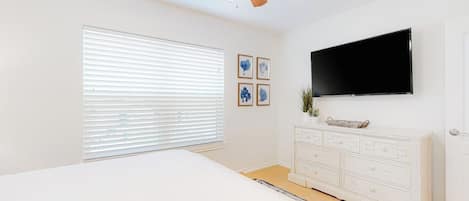 Un dormitorio con cama, ventilador de techo, televisor montado en la pared y cómoda blanca.