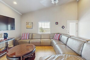 A living room with a sectional sofa, a coffee table, a flat-screen TV, and a window with blinds. - Casa Nirvana - Steps to the Beach & Balcony (South Padre Island)