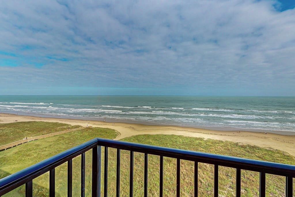 A coastal view from a balcony with a railing, overlooking a beach and ocean.