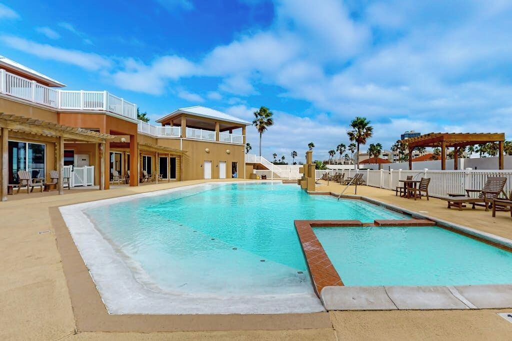 A swimming pool with lounge chairs and a covered area, surrounded by buildings with balconies and palm trees.