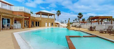 A swimming pool with lounge chairs and a covered area, surrounded by buildings with balconies and palm trees.