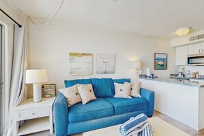 Ein Wohnzimmer mit blauem Sofa, einem weißen Beistelltisch mit Lampe sowie einer Küchenzeile mit Arbeitsplatte und Schränken.