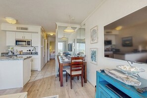 Cozinha moderna com armários brancos, área de refeições com mesa e cadeiras de madeira, e um rack azul com televisão.