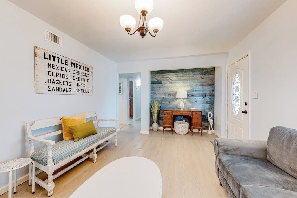 A living room with a white sofa, a bench with cushions, a fireplace, and a wall-mounted sign listing items like 'MEXICAN DRESSES' and 'CURIOS CERAMIC'.