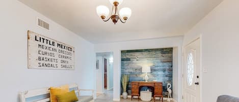 A living room with a white sofa, a bench with cushions, a fireplace, and a wall-mounted sign listing items like 'MEXICAN DRESSES' and 'CURIOS CERAMIC'.