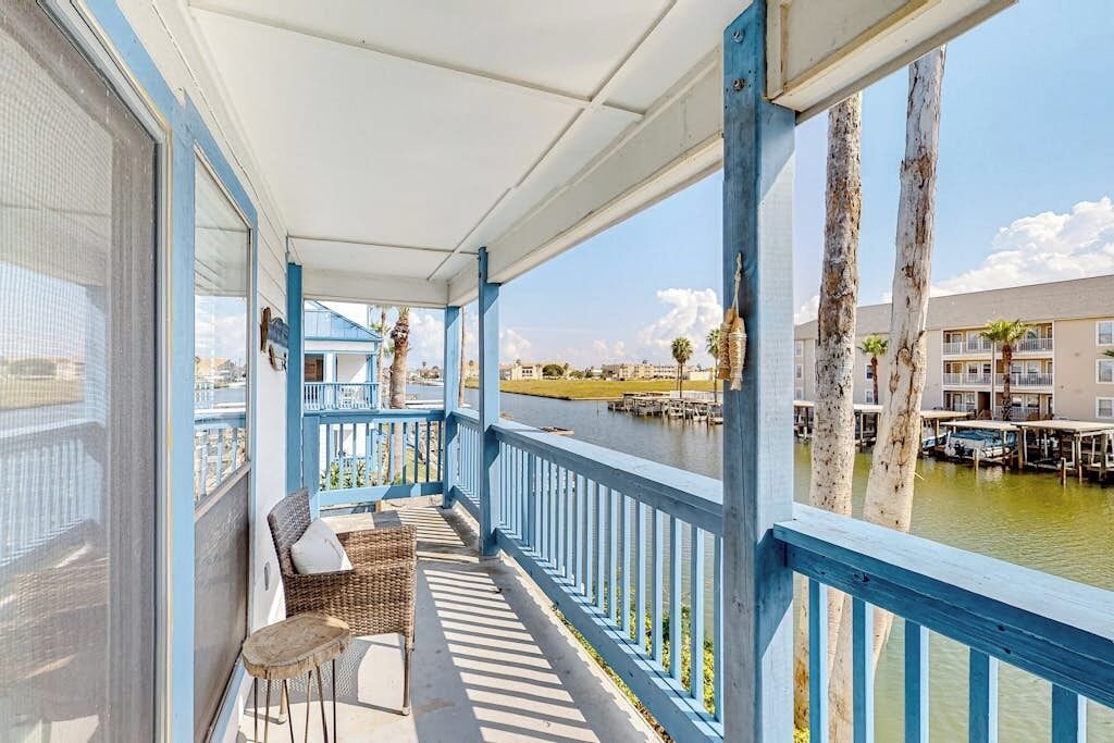 Een overdekt balkon met rieten meubilair, met uitzicht op een waterpartij en gebouwen.
