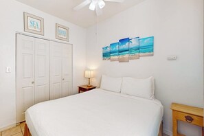 Ein Schlafzimmer mit einem Bett, einem Nachttisch mit Lampe, einem Deckenventilator und Wandbildern, die eine Strandansicht zeigen.
