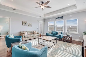 A spacious living room with a ceiling fan, two sofas, a coffee table, and a large window with blinds. - The Blue Shell - Pool, Big Deck & Dog-Friendly (Port Aransas)