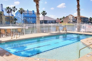 A swimming pool with a white fence, palm trees, and buildings in the background. - Compass Condominiums A7 (Corpus Christi)