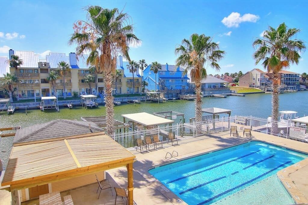 A swimming pool with a wooden deck and lounge chairs, surrounded by palm trees and a marina with boats.