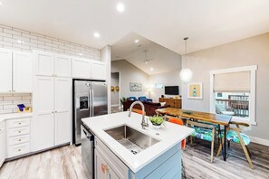 A modern kitchen with white cabinets, a stainless steel refrigerator, and a dining area with a wooden table and chairs. - Martinis & Marlin - Heated Pool (Port Aransas)