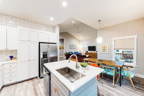 A modern kitchen with white cabinets, a stainless steel refrigerator, and a dining area with a wooden table and chairs. - Martinis & Marlin - Heated Pool (Port Aransas)