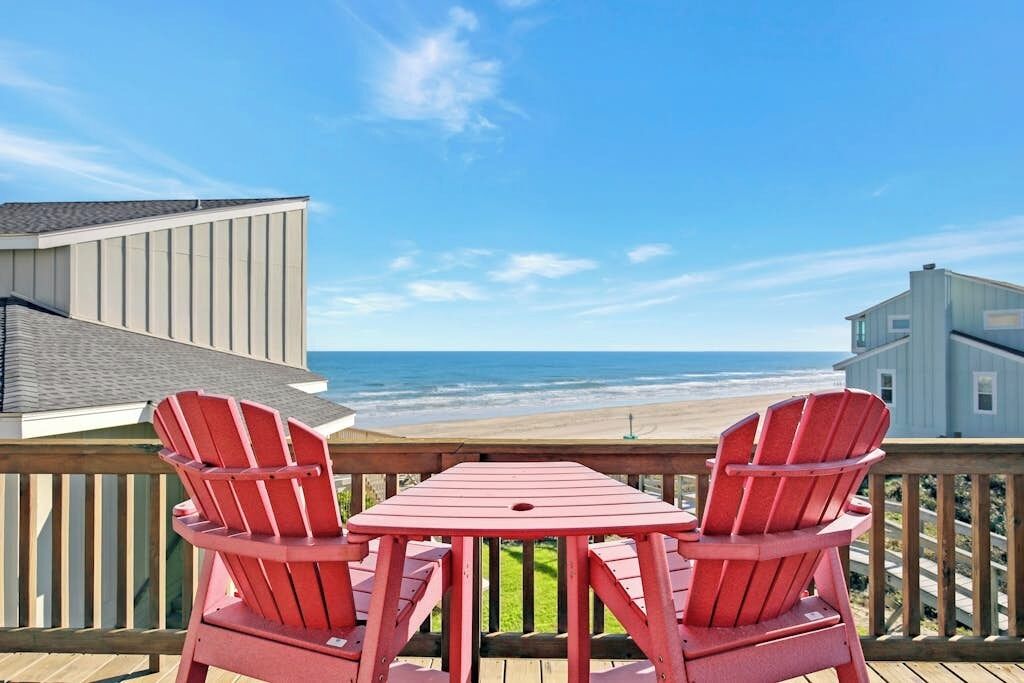 A deck with two red Adirondack chairs and a table overlooking a beach.