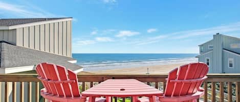 A deck with two red Adirondack chairs and a table overlooking a beach.