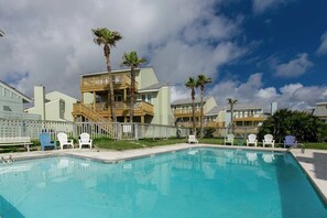 Ein Pool mit Holzterrasse, umgeben von Liegestühlen und Palmen, mit einem mehrstöckigen Gebäude im Hintergrund.