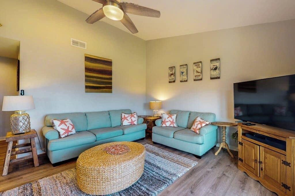 A living room with two sofas, a television, a ceiling fan, and a woven ottoman.