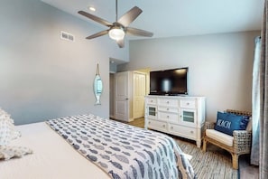A hotel room with a bed, a television, a ceiling fan, a white dresser, and a wicker chair. - Beachaholic - Bay Views, Pool, Near the Beach (Rockport)