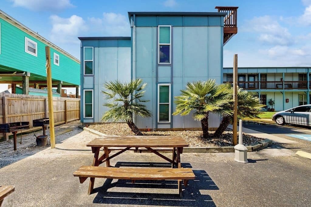 Un bâtiment à la façade bleue, une table de pique-nique en bois au premier plan et une voiture garée à l’arrière-plan.