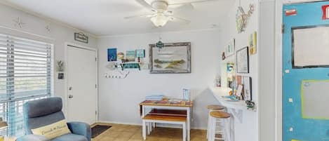 A room with a ceiling fan, a desk with a chair, a small table with a bench, and a door with artwork.
