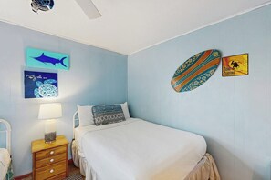 Ein Schlafzimmer mit einem Bett, einem Nachttisch, einer Lampe und Wandkunst, darunter eine Haifischsilhouette, ein blaues abstraktes Design und ein Surfbrett mit der Aufschrift 'Gone Surfing'.