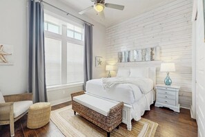 Um quarto com cama, mesa de cabeceira, cadeira, banco e janela com cortinas.