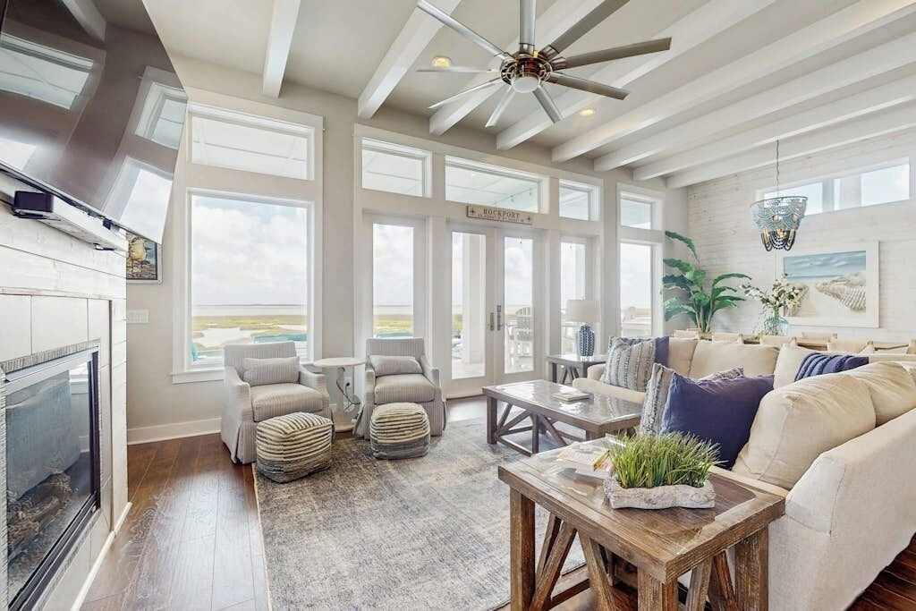A spacious living room with a fireplace, large windows, and a ceiling fan.