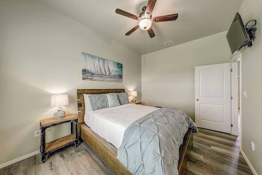 A bedroom with a bed, bedside tables, a ceiling fan, a painting, and a door.