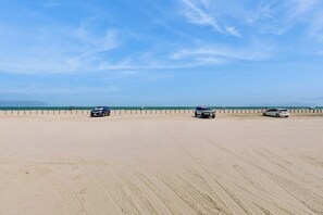 Ein Sandstrand mit drei geparkten Fahrzeugen am Ufer, einem klaren Himmel und dem Ozean im Hintergrund.