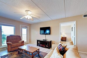 A living room with a ceiling fan, a flat-screen TV, a sofa, a coffee table, and a window with a view. - Suntide II Condominiums #308 (South Padre Island)