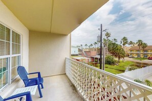 A balcony with a white lattice railing, a pair of blue chairs, and a view of a residential area with palm trees and a body of water. - Fiesta Sol Condominiums 211 (South Padre Island)