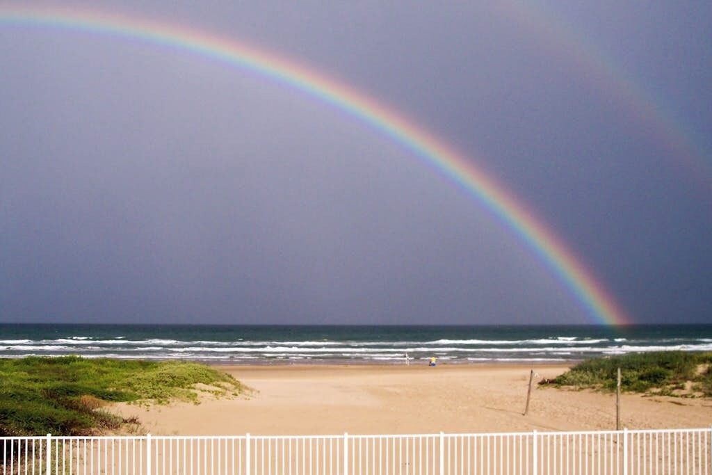 Une plage avec une clôture blanche et un arc-en-ciel dans le ciel.