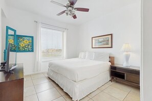 A hotel room with a bed, a desk, a chair, a ceiling fan, a window with blinds, and artwork on the walls. - Las Marinas Condominiums 405 (South Padre Island)