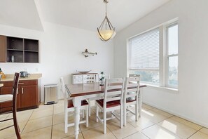 Un coin repas avec une table en bois et des chaises blanches, un luminaire suspendu et une fenêtre avec des stores.