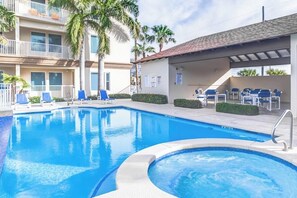 Piscina com espreguiçadeiras e banheira de hidromassagem, cercada por palmeiras e um edifício com sacadas.