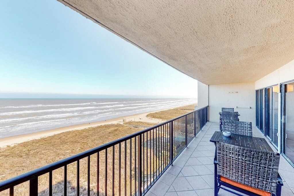 A balcony with a view of the beach, featuring a railing, two wicker tables, and a bench.