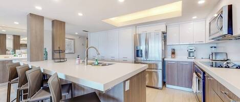 Una cocina moderna con una isla central, taburetes de bar, electrodomésticos de acero inoxidable y armarios blancos.