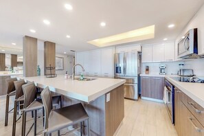A modern kitchen with a central island, bar stools, stainless steel appliances, and white cabinetry. - Ocean Vista Condominiums #1004 (South Padre Island)