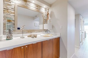 A bathroom with a white countertop, a wooden cabinet, a large mirror, and decorative items including a jar of stones and a bird sculpture. - Ocean Vista Condominiums #1004 (South Padre Island)