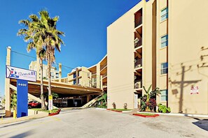 A multi-story hotel with a parking garage entrance, palm trees, and a clear blue sky. - Suntide II Condominiums #107 (South Padre Island)