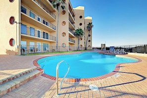 A swimming pool with a brick border, surrounded by a building with balconies and palm trees. - Suntide II Condominiums #107 (South Padre Island)