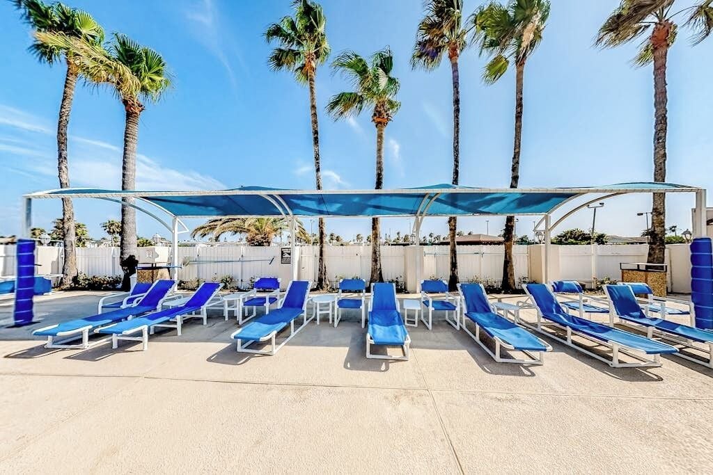 A poolside area with sun loungers and a covered seating area, surrounded by palm trees.