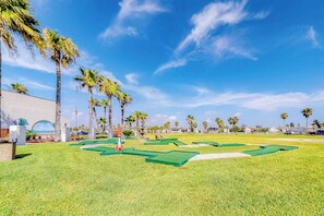 A golf course with a clear sky, palm trees, and a building with a large arched entrance. - Long Island Village 349 Sand Dollar (Port Isabel)