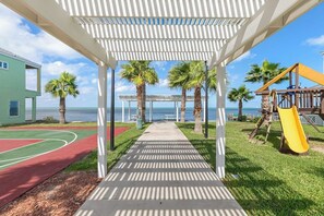 A covered walkway leading to a beach area with a playground and a basketball court. - Las Joyas Bayview Home #216 (Port Isabel)