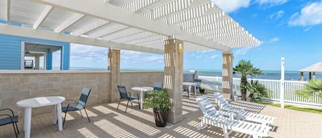 A covered outdoor area with tables and chairs, a view of the ocean, and a building with a blue exterior.