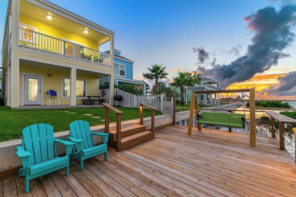 A two-story house with a deck, outdoor seating, and a view of the ocean at sunset.