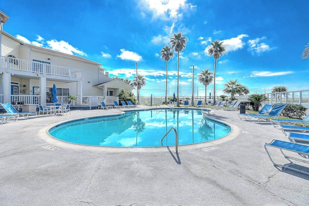 A pool area with lounge chairs and palm trees, a building with a balcony, and a clear sky.