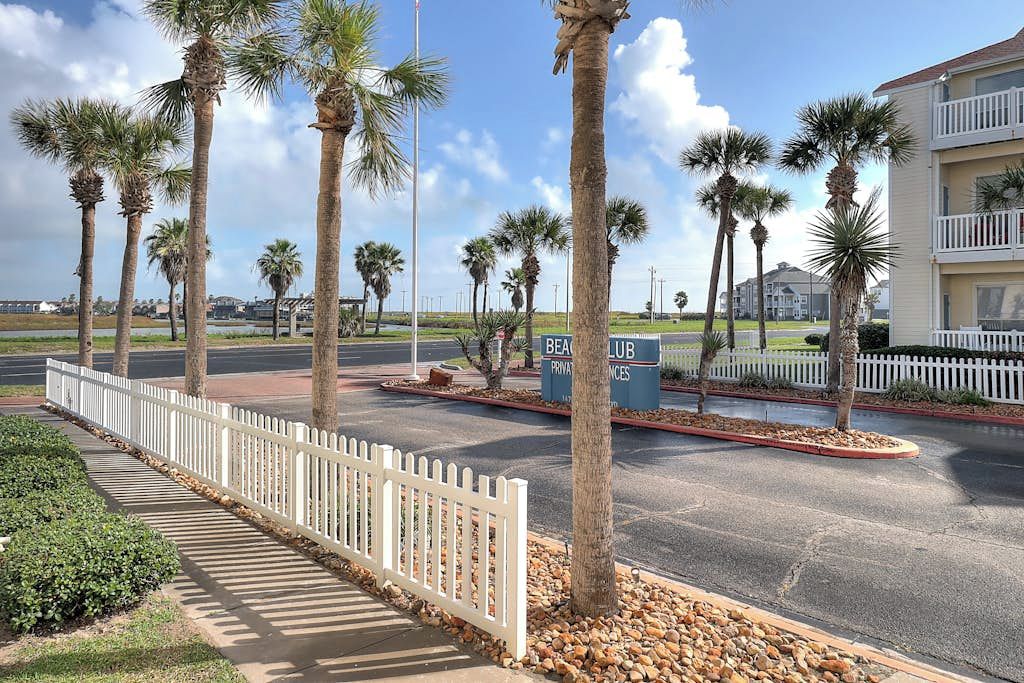 A gated entrance with palm trees, a white picket fence, and a sign for a beach club.