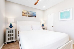 A bedroom with a large bed, two bedside tables with lamps, a ceiling fan, and artwork on the walls. - Beach Club 173 & 174 (Corpus Christi)