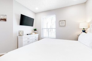 Een slaapkamer met een bed, een aan de muur bevestigde televisie, een raam met zonwering, een nachtkastje met een lamp en een dressoir met decoratieve objecten.